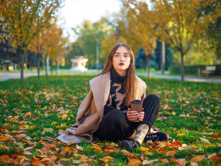 Girl in a beige coat on the background of the autumn park and yellowed leaves on the tree, thoughtfully sits on a green meadow, fashion portrait young womanの写真素材