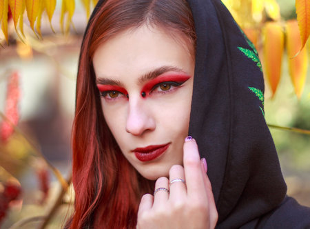 Mystical portrait of stylish red-haired girl in black coat on the background of colorful trees in...