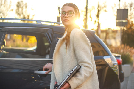 Young confident luxurious young business lady in white coat gets out of passenger seat in car with...