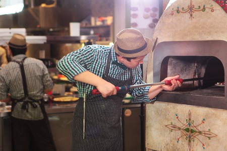 A young guy, an Asian-looking pizza maker places the finished pizza in a big and beautiful oven. Pizza on a big shovel. The interior of the restaurant or cafe.の写真素材