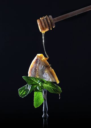 A drop of honey flows to a piece of cheese. A piece of hard cheese on a vintage fork on a black background. Photo on a black background. Isolated object.の写真素材
