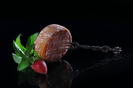An antique fork is stuck in a piece of cheese. A round piece of cheese and strawberries on a black background. Minimalism and macro photography. Copy spaceの写真素材