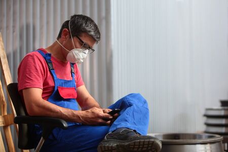 A man in a mask at the factory while resting holds a phone in his hands. The concept of workforce at the factory.の写真素材