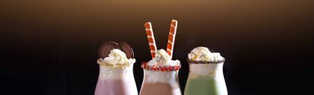 Three milkshakes on a black background. decorated with cream, wafer rolls and cookies. Long banner....
