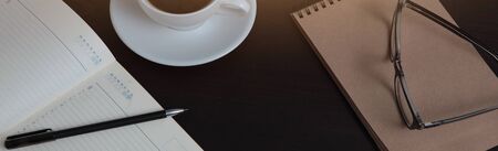 An open notebook and eyeglasses on the table. A white Cup of coffee. Long banner. The view from the...