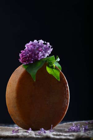 Round head of hard cheese with a branch of lilac on a black background. Vertical photo. Copy space.の写真素材
