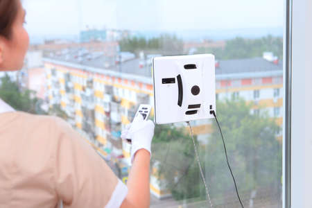 A girl in uniform remotely controls a robot that cleans Windows. View from the window. Concept of cleaning in a hotel or at home. Cleaning of hotel rooms. The man is out of focus. Unrecognizable photo.Back viewの写真素材