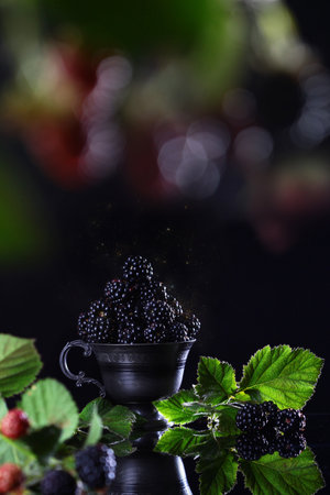 BlackBerry berry with leaves in a black metal Cup on a black background. Copy of the space. The concept of still life. Summer season. Vertical photo. Berries out of focus bokehの写真素材