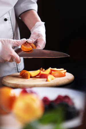 The cook cuts the fruit into slices. Assorted fruits and berries are cut into pieces. Unrecognizable vertical photo. Only hands with gloves.の写真素材