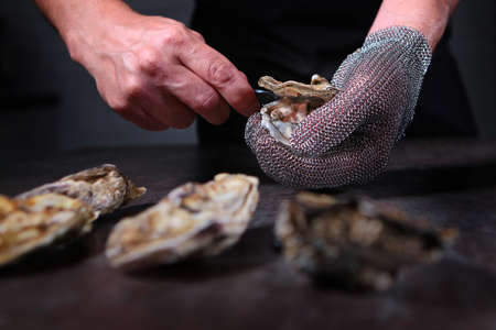 A man in protective gloves opens an oyster shell. Protein delicatessen food. Dark background. Unrecognizable photo. Fresh seafood.の写真素材