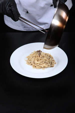 The chef puts the pasta with truffle sauce from the pan on a plate. Pasta with black truffle. Unrecognizable person. Photo on a black background. Copy space. Vertical photo. Top view.の写真素材