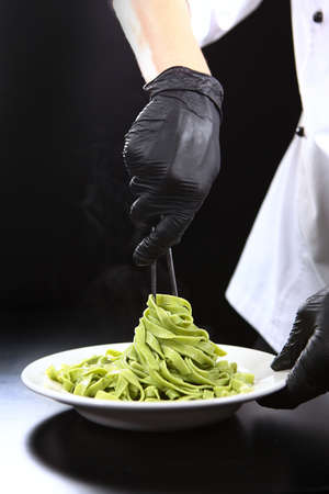 The chef uses tweezers to spread the freshly prepared spinach paste on a white plate. The concept of restaurant or home cooking.An unrecognizable man in protective gloves. Photo on a black background. Vertical photo.の写真素材
