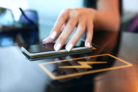 Wireless charging of your phone or gadget in a public place or in a cafe. A woman's hand holds the phone and charges the gadget wirelessly. Unrecognizable person. Dark background. Copy space. Indoors.の写真素材