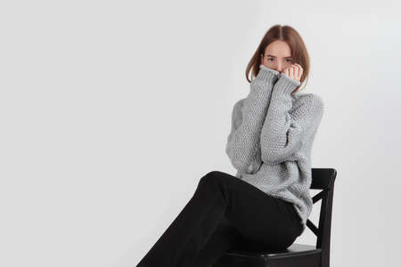 A girl warms her frozen hands in a woolen sweater. A girl in black trousers sits on a black chair. Natural wool clothing for the cold season. Light background. Copy space.の写真素材