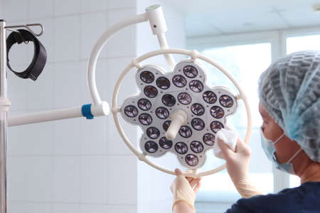 Disinfection of medical equipment in the clinic. An employee of a dental clinic wearing protective gloves and a medical mask wipes the equipment in surgical room. LED lamp in the surgical room. Rear view.の写真素材