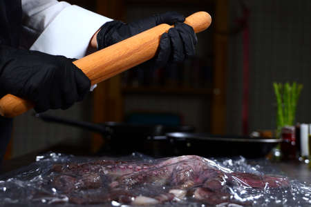 The cook is holding a wooden rolling pin. Octopus in plastic wrap. Seafood dishes. Protein food. An unrecognizable person. Side view.の写真素材