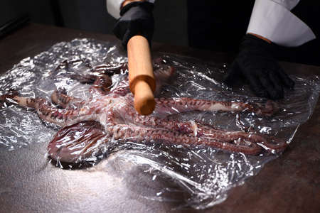 The chef uses a wooden rolling pin to beat up a fresh whole octopus. Octopus in plastic wrap. Seafood dishes. Protein food. An unrecognizable person. Top view. The cook's hands are covered with protective gloves.の写真素材