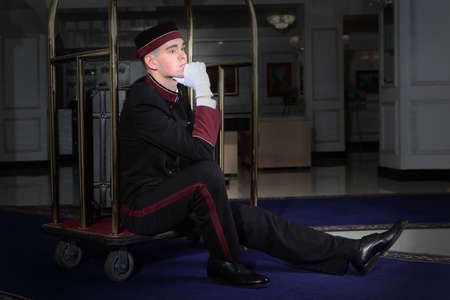 A pensive or tired uniformed doorman sits on a luggage trolley. Hands in white cotton gloves. Restaurant business concept.の写真素材