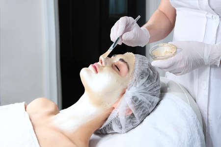 Cosmetologist uses a brush to apply a gold-colored cosmetic mask to a woman's face. The concept of beauty and preservation of youth. The procedure in a cosmetology clinic.の写真素材