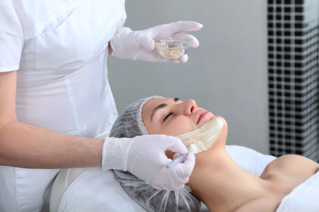 Applying a mask to the face of a young woman in a cosmetology clinic. The concept of preserving youth and caring for the skin of the face. Gold-colored alginate mask.Side view. An unrecognizable person.の写真素材