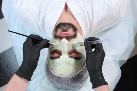 Beautician applies a nourishing mask to the mans face. A man doing facial procedures in a cosmetology clinic. Mens cosmetology. The concept of preservation of youth and skin care of the face.Top view.の写真素材