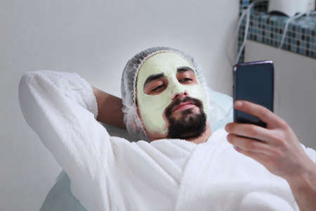 Cosmetic nourishing mask for a mans face. A man does facial procedures in a cosmetic clinic and holds a phone in his hand. Mens cosmetology. The concept of preservation of youth and skin care of face.の写真素材