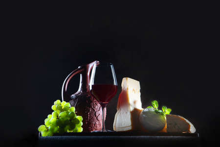 Assorted cheeses and homemade red wine. Hard and soft cheeses. Homemade wine in a ceramic jug. Freespace . Photo on black background.Green grapesの写真素材