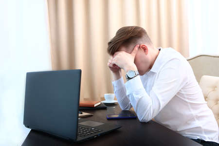 A frustrated young businessman stands in front of a desk with a laptop. He put a hand to his head....