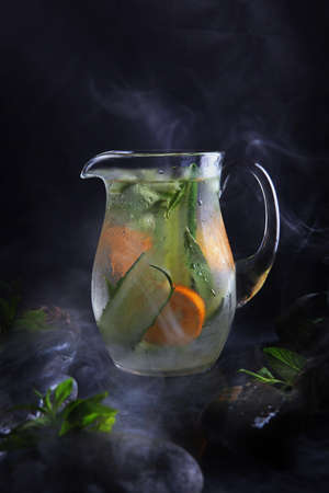 Homemade cold lemonade based on cucumber, mint and orange. Fresh fruit in a misted glass jar. Black background. Vertical photo. Wet stones and smoke.の写真素材