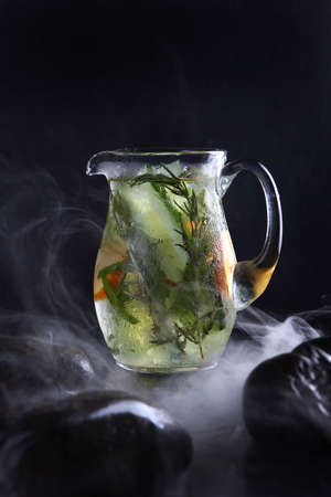 Homemade cold lemonade based on orange, cucumber and rosemary. Fresh fruits and vegetables in a misted glass jar. Pieces of ice. Black background. Vertical photo. Refreshing drink.Quench your thirst.の写真素材