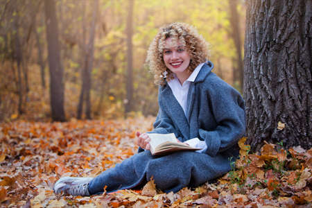 Beautiful girl reads a book in autumn park. Girl has curly hair. Book in hand. Girl is dressed in...