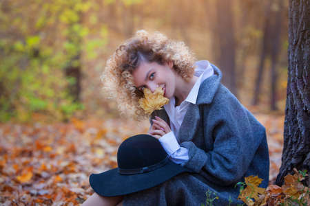 A young girl sits in a park in autumn. Girl smiles and holds an autumn maple leaf in her hand. Warm clothes. Yellow autumn leaves. Copy space. Blurred background.の写真素材
