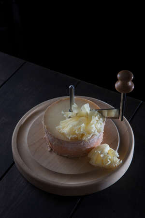 A Swiss semi-hard cheese made from unpasteurized cow's milk called a monk's head. The cheese stands on a wooden round board. Special girolle knife.With copy space. Vertical photo on a black background.の写真素材