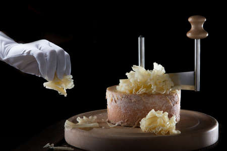 A hand in a white glove holds thin slices of tete de moine cheese. Thin shavings of Swiss elite tete de moine cheese. Special girolle knife. Black background.の写真素材