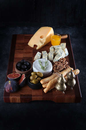 Several types of cheese on a wooden board with smoked sausage and olives. Figs and grissini. Honey and cheese on a wooden board. selective focus. dark background. vertical photo.の写真素材