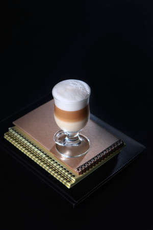 A coffee drink in a tall glass sits on a stack of notebooks. A three-layer drink made of coffee, milk and creamy foam.Concept of a business breakfast or coffee break. Food and drinks.の写真素材