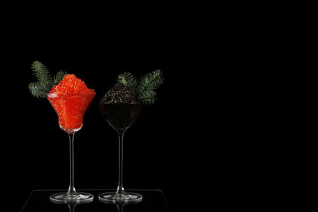 Caviar is black and red in tall glasses. The wine glasses are decorated with fir branches. A festive snack.Salmon caviar. Delicacies.Space for text. Black background. New Year or Christmas.の写真素材