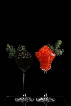 Caviar is black and red in tall glasses. The wine glasses are decorated with fir branches. Festive snack.Salmon caviar. Space for text. Black background. New Year or Christmas. Vertical photo.の写真素材