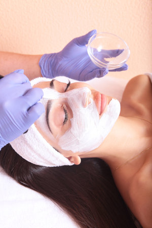 Beauty procedure in a beauty salon. Cosmetic mask on the girls face. The cosmetologist puts a mask on the face of a young girl. The concept of beauty and facial skin care. Vertical photo.の写真素材
