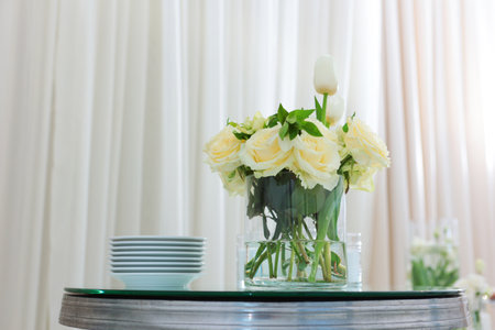 On the table there is a vase with white roses and a stack of plates. Set table at a party. Mirrored tabletop.The concept of public catering.の写真素材
