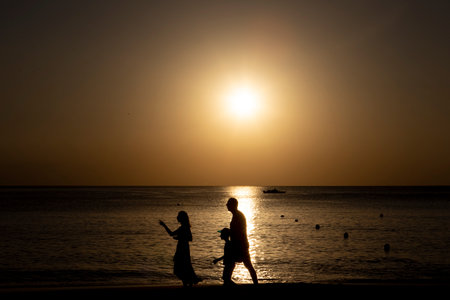 The black silhouette of a happy family against the background of the sun. Father, mother and son are walking along the ocean shore. Lifestyle travel, parentsの写真素材