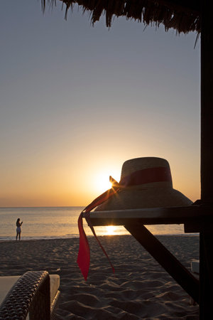 Womans hat with ribbon on beach. Dawn on coast of Indian Ocean. An unrecognizable female figure in background. Concept of leisure and travel.の写真素材