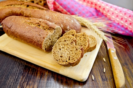 Rye baguettes on a board with a knife, wicker basket, stalks of rye, a napkin on a dark wooden board