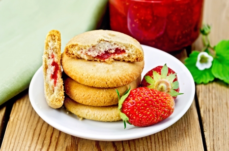 A stack of cookies filled with jam and strawberries on a plate, a jar of strawberry jam on the background of wooden boardsの写真素材