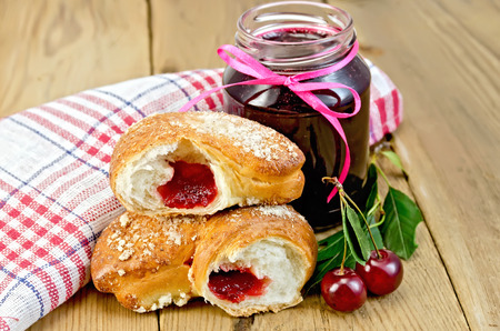 Puff bun with jam, a jar of cherry jam, cherries with leaves, a napkin on a wooden boards backgroundの写真素材