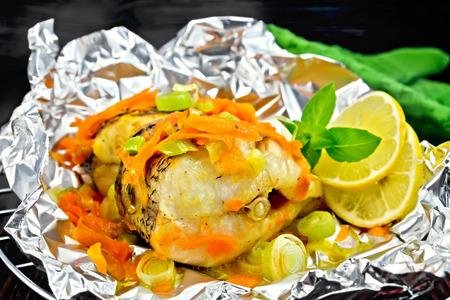 Pike with carrots, leek, basil and lemon slices in a foil on a metal grid, green towel on a dark wooden boardの写真素材