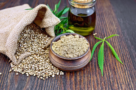Hemp flour in a clay bowl, the grain in the bag and on the table, the oil in a glass jar, leaves and stalks of cannabis on a background of dark wooden boardsの写真素材