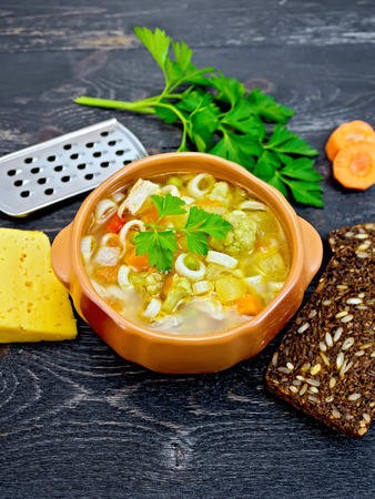 Minestrone soup with meat, celery, tomatoes, zucchini and cabbage, green peas, carrots and pasta in an earthenware bowl, cheese, grater on black background wooden plankの写真素材