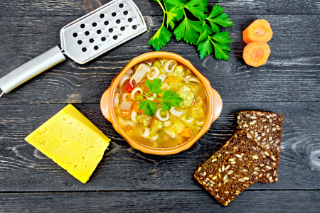 Minestrone soup with meat, celery, tomatoes, zucchini and cabbage, green peas, carrots and pasta in an earthenware bowl, cheese, grater on black background wooden plank on topの写真素材