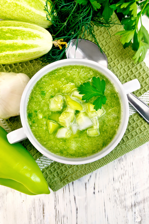 Cucumber soup with green peppers and garlic in a white bowl on a napkin, parsley on a background of wooden boards on topの写真素材
